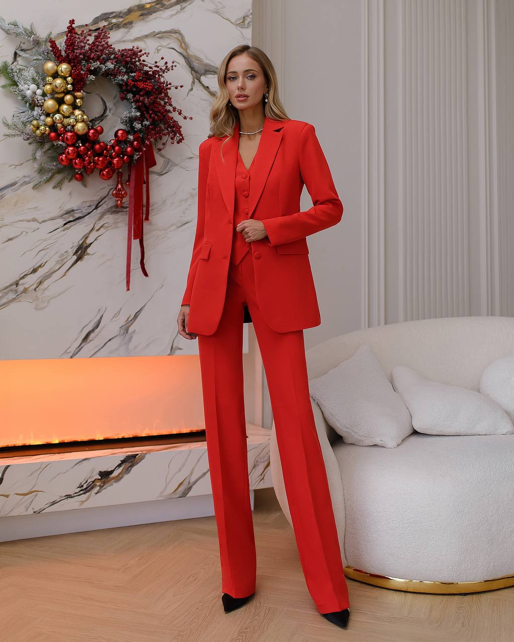 a woman in a red suit stands in front of a christmas wreath