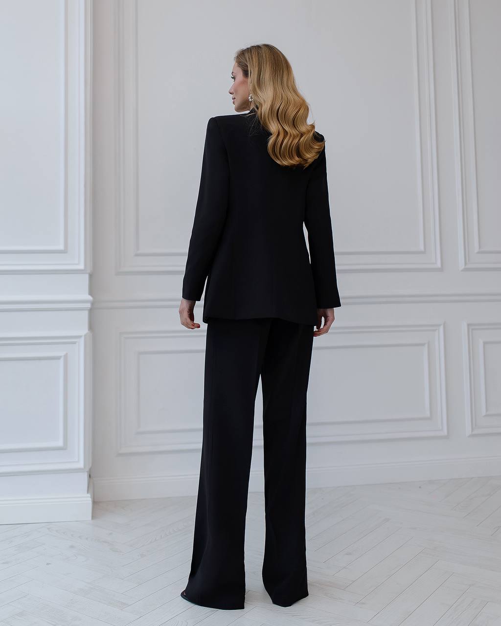 a woman in a black suit stands in front of a white wall